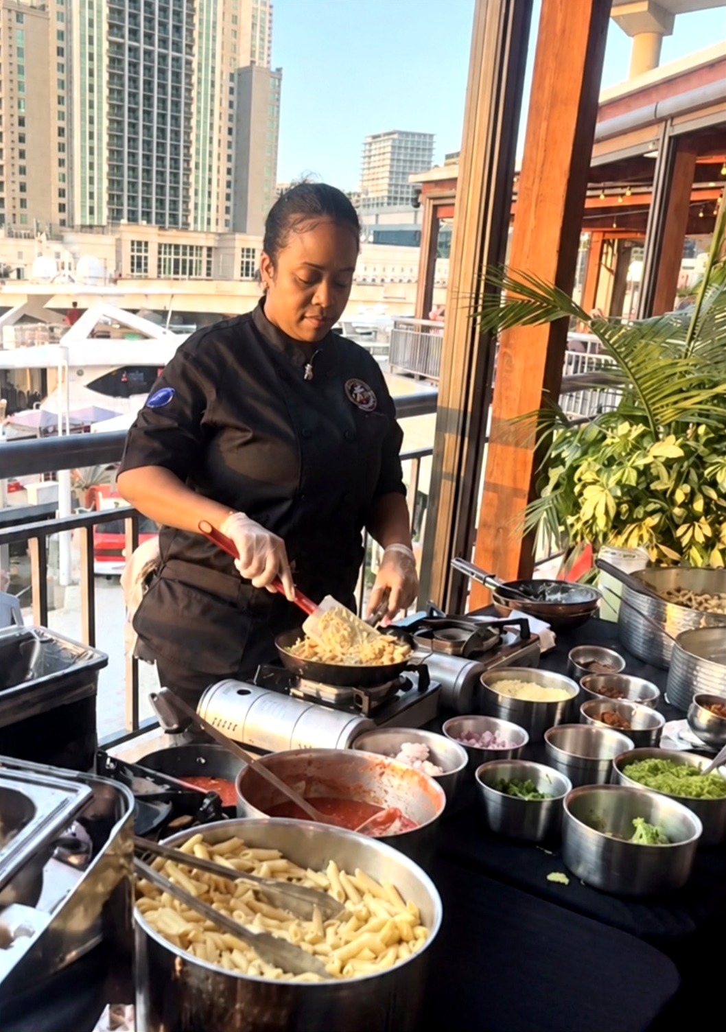 Chef Candy tossing penne at an outdoor catering station with a waterfront skyline behind her
