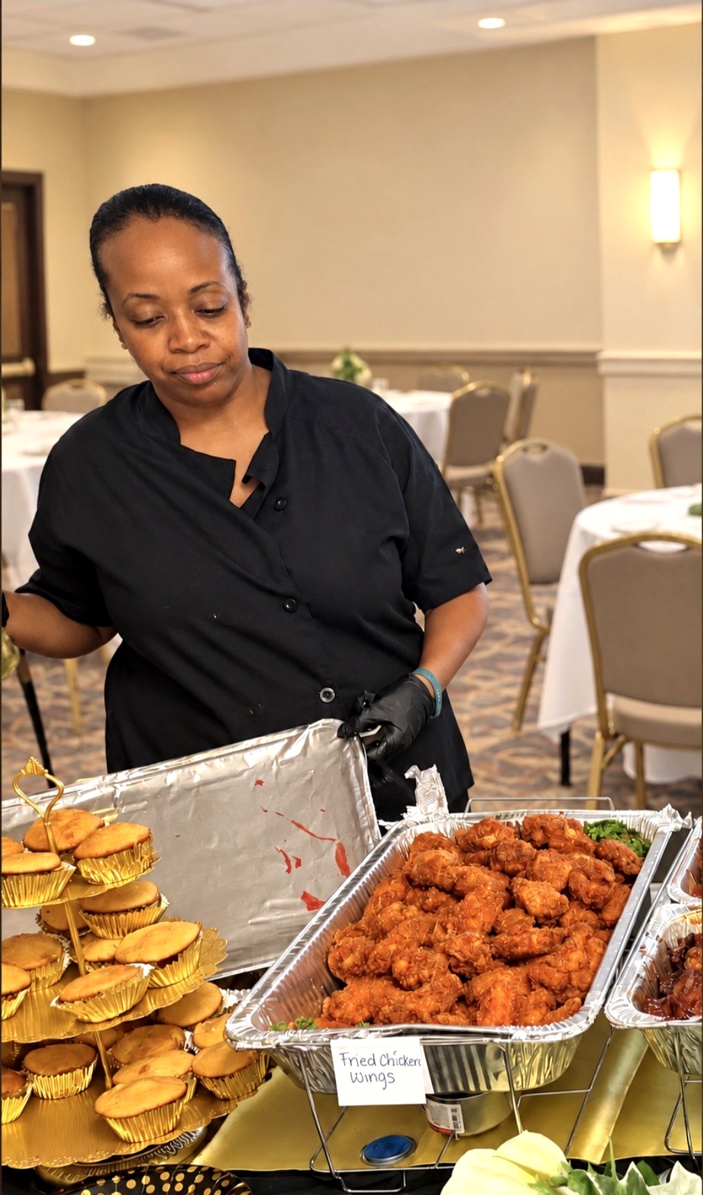 Chef Candy serving fried chicken wings from a labeled chafing dish at a full-service catered event, with a tiered gold stand of cornbread muffins beside her
