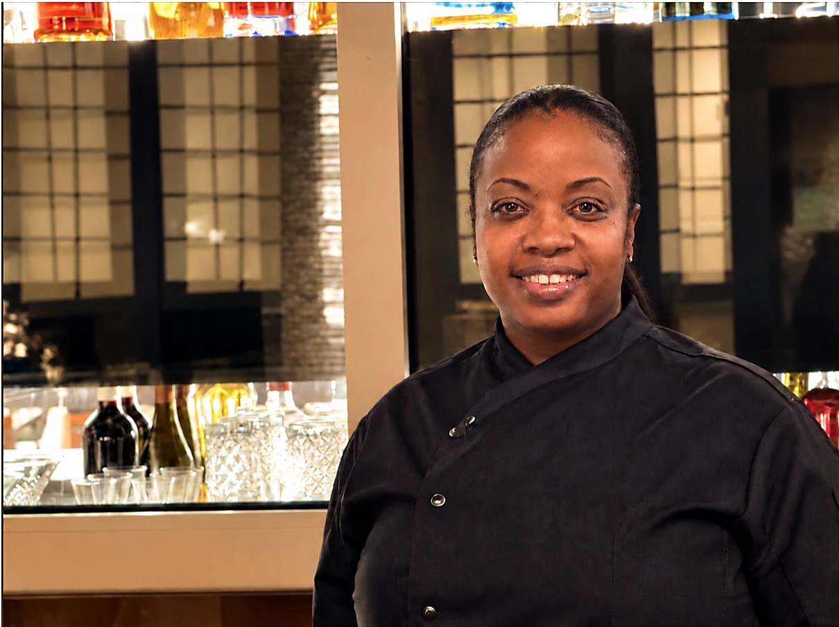 Chef Candy smiling in her black chef jacket in front of a bar